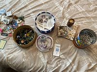 Full lot laid out on a bed: miniature tea sets, wicker basket with marbles, container with small marbles or stones, box of seashells, and colorful keychains scattered