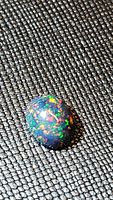 Single natural black opal oval cabochon gemstone pictured on textured dark fabric background showcasing its multicolored play-of-color effects.