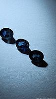 Three oval blue sapphire gemstones arranged in a row on a textured white surface, showing their shape, color, and faceting.