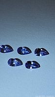 Five pear cut natural Tanzanite gemstones in calibrated sizes laid out on a flat surface, showing purple blue color and good clarity.