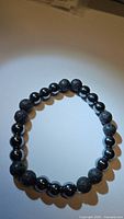Top-down image of round beaded bracelet with alternating black porous lavastone beads and smooth shiny hematite beads on elastic cord, displaying full circular shape on neutral background.