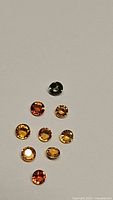 Photo shows nine round faceted natural rainbow sapphire gemstones in different colors including orange, yellow, and dark greenish-blue, arranged on a plain background.