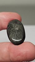Close-up photo of the natural silver sheen obsidian oval cabochon held between thumb and forefinger, showing its polished surface and silver shimmer effect.