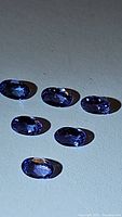 Photo showing six oval cut blue tanzanite gemstones placed on a flat surface, highlighting their deep blue color and faceted cuts.