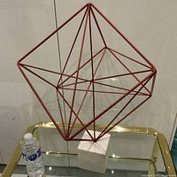 Front angled view of the large metal geometric cube sculpture on white marble base, showing overall size and structure with water bottle for scale.