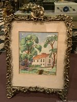 Close-up photo of the framed watercolour painting showing the ornate detailing of the frame and the artwork featuring a house, trees, and greenery.