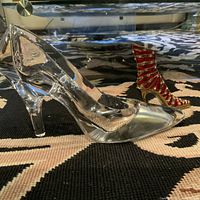Large clear glass high-heeled shoe with smaller red enamel and metal boot shoe decor pieces side by side on patterned fabric surface.