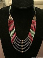 Front view of the five-strand necklace displayed on a black velvet bust stand showing the arrangement of red ruby, light green emerald, and dark blue sapphire beads separated by silver spacers and silver fittings.