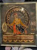 Front view of the vintage enamel on metal folklore picture showing the female figure seated with spinning wheel and stool, framed in metal.