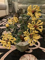 Front view of the full plant showing multiple yellow glass flowers with curled petals and green glass leaves in a ceramic pot on carpet.