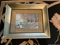Framed vintage enamel painting on copper lying on a patterned textile, showing overall artwork and frame detail.