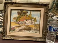 Frontal photo of vintage oil painting in decorative frame showing a house in a rural setting with a stone bridge. The frame shows wear and damage.