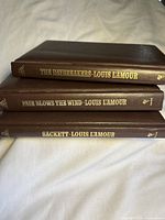 Three brown leather bound Louis L'Amour books stacked showing the spines with gold embossed titles