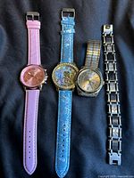Full view of the three watches and the bracelet laid on dark fabric, showing different strap materials and styles including pink leather, blue embossed leather, expandable metal band, and stainless steel bracelet with black highlights