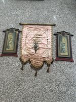 Three antique Asian wall decor pieces laid flat on carpet: two wooden plaques with painted figures and one pink silk embroidered banner with tassels, showing overall condition and appearance.
