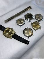 Six vintage watch heads and one spare metal band laid on white fabric, varying dial colors and styles visible