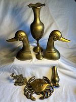 Seven brass decorative items including two tall etched vases with flared tops, a pair of duck bookends, a moose figurine, crab door knocker, and small vase displayed on white fabric.