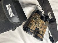 Bushnell branded camouflage binoculars with neck strap and carrying case shown from a side angle.