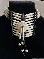 Full front view of the indigenous bone bead choker necklace on black background, showing rows of white tubular beads and round dark beads with leather backing and strap closure.