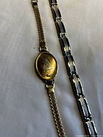 Two women's bracelets shown side by side on a white surface, one gold-tone with an engraved girl on an oval plate and mesh chain band, the other silver-tone with black rectangular linked design with silver beaded accents.