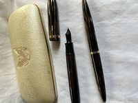 Photo of the fountain pen and mechanical pencil separated from their caps alongside the original cream textured carrying case with embossed emblem.