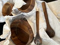 Two wooden salad serving utensils - one fork and one spoon - alongside some wooden bowls wrapped in white paper.