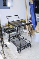 Side view of black wrought iron serving cart showing two mesh shelves and scrollwork design on the sides. Cart placed in garage area with some building materials and buckets in the background.