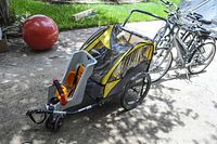 Baby bike carrier attached to a bicycle outdoors, showing the black and yellow weather canopy, two large wheels, tow bar, and integrated baby seat.