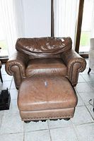 Brown leather oversized arm chair with removable seat cushion and matching ottoman, both showing nailhead trim details and bun feet on chair.