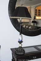 Front view of vintage style lamp with black fabric shade with gold trim on a decorative cast ceramic base, sitting on a dark wooden table.