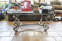 Full frontal view of the glass topped coffee table showing the rectangular clear glass and metal curled scroll base under natural light with some items on top.