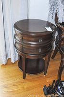 Front view of the round side table showing the two stacked curved drawers closed, dark wood finish, standing on hardwood floor next to a chair.
