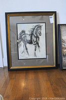 Full view of framed horse artwork in black and gold frame with beige matting.
