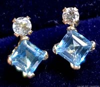 Close-up photo showing one pair of square-cut blue topaz earrings accented with round diamonds on 10K yellow gold settings against a dark blue background.