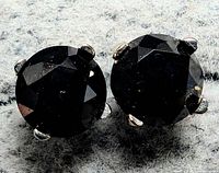 Close-up photo of 14K white gold black diamond stud earrings on gray fabric surface, showing round faceted black diamonds set in four-prong white gold mountings.