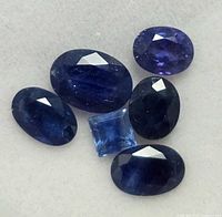 Close-up image showing six blue sapphire gemstones cut in oval and square shapes on a light-colored background.