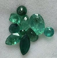 Multiple loose emerald gemstones of various cuts and shades lying on a light grey textured background. The stones include oval, round, marquise, and pear shapes with visible natural inclusions.
