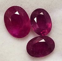 Three oval-shaped, faceted genuine enhanced ruby gemstones placed closely together on a white background, showing color and clarity.