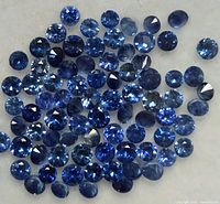 Multiple small loose round faceted blue sapphire gemstones spread on a white surface showing various sizes and blue color shades.