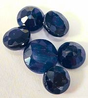 Seven loose round and oval faceted blue sapphires arranged on a flat surface with visible facets and deep blue color.