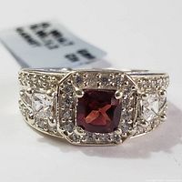 Close-up image of a silver garnet ring showing cushion-cut garnet, clear stones on sides, and small clear stones on band