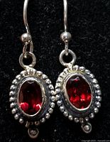 Close-up photo of two silver earrings showing dark red oval garnet stones with beaded silver border and hook fastening on black background.
