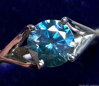 Close-up photo showing the round, blue moissanite gemstone in a four-prong silver setting.