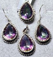 Front view showing all four pieces: ring, pendant, and two earrings laid on grey fabric background. Highlights the pear-shaped mystic topaz stones and silver settings.