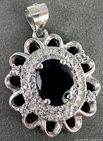 Close-up photo of silver pendant with dark blue oval sapphire center set in silver with surrounding rows of small clear stones and decorative scalloped border.
