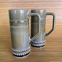 Two tall ceramic mugs or beer steins with angular handles, grey-green glaze with textured horizontal bands near top and base
