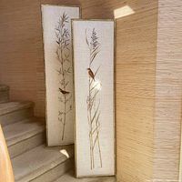 Full view of two tall framed needlepoint panels leaning on stairs, showing birds and botanical designs.