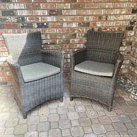 Two outdoor wicker chairs with light gray cushions against a brick wall