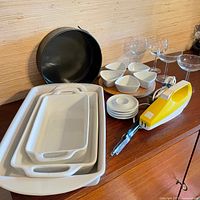 Overview of all items including three white ceramic rectangular baking dishes stacked, one black round baking pan behind, six white triangular bowls, four small white plates stacked, four clear stemmed glasses, and one yellow hand appliance with metallic tool.