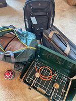 Overview of lot showing camping stove, fuel canisters, sleeping bag, duffel bag, and two soft carrying bags including black luggage and brown case.